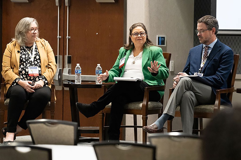 Three people sitting in high top chairs addressing an audience in a conference setting.