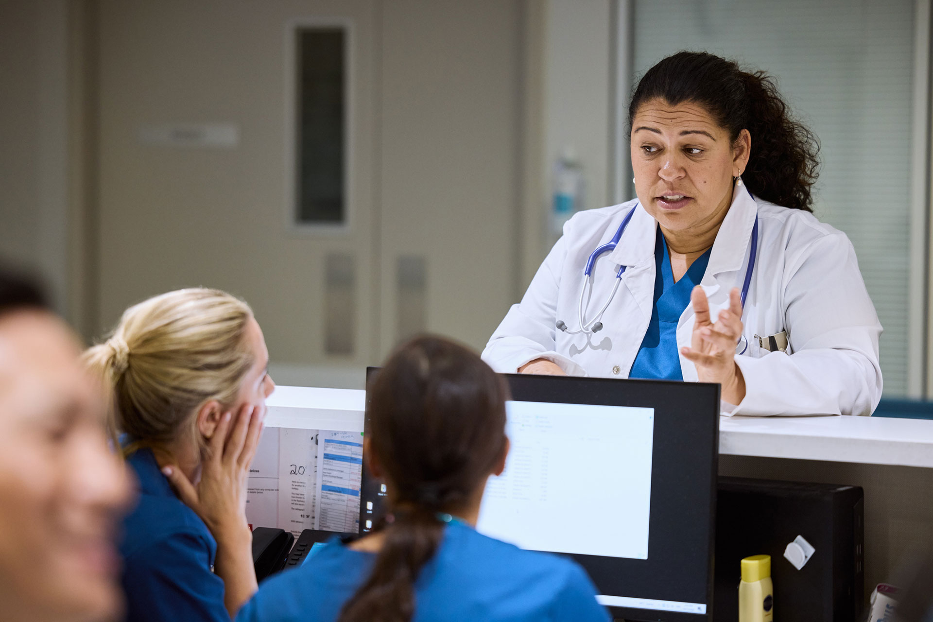 Female doctor talking to nurses at hospital reception. Medical team is discussing patient care while reviewing information on computer. Healthcare professionals are working together for better treatment.
